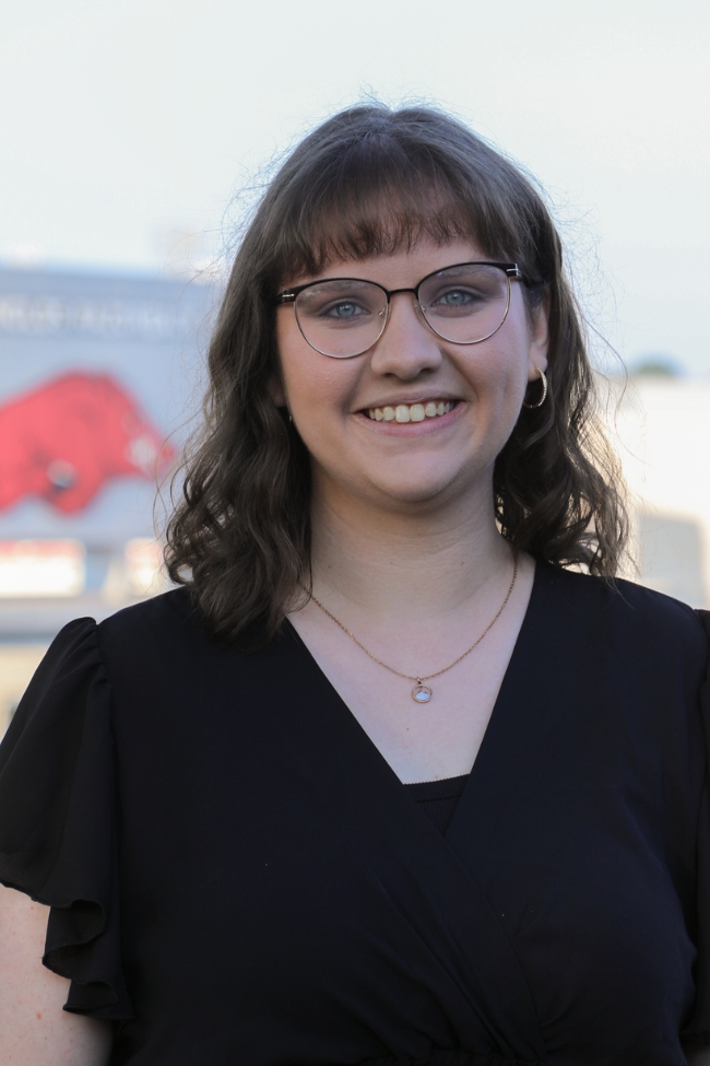 Professional headshot of a (Natasha Hightower) young woman with brown hair and glasses, wearing a black top. The University of Arkansas Razorback logo is visible in the blurred background.